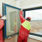 Two construction workers in red uniforms install a window.