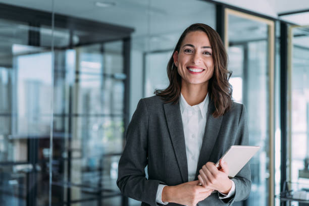 A confident businesswoman stands in a modern office, holding a tablet and smiling, representing compliance in UAE business.