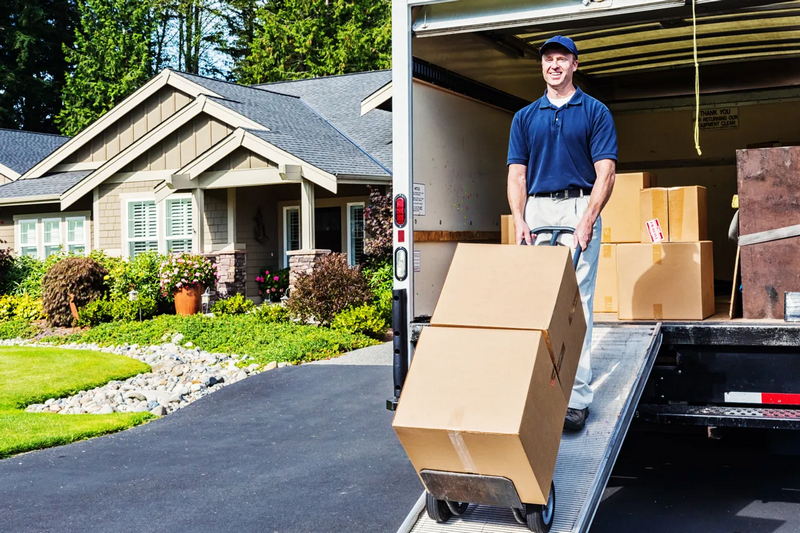 A moving company worker unloads boxes from a truck in front of a suburban house.