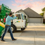 A person is wheeling a green trash bin towards a waste management truck on a suburban street at sunset.