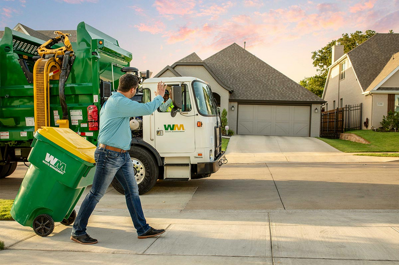 A person is wheeling a green trash bin towards a waste management truck on a suburban street at sunset.