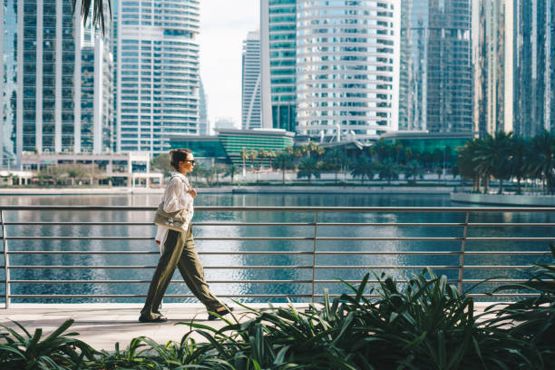 A woman walks along a waterfront in Dubai, showcasing the city's modern skyscrapers and urban lifestyle.